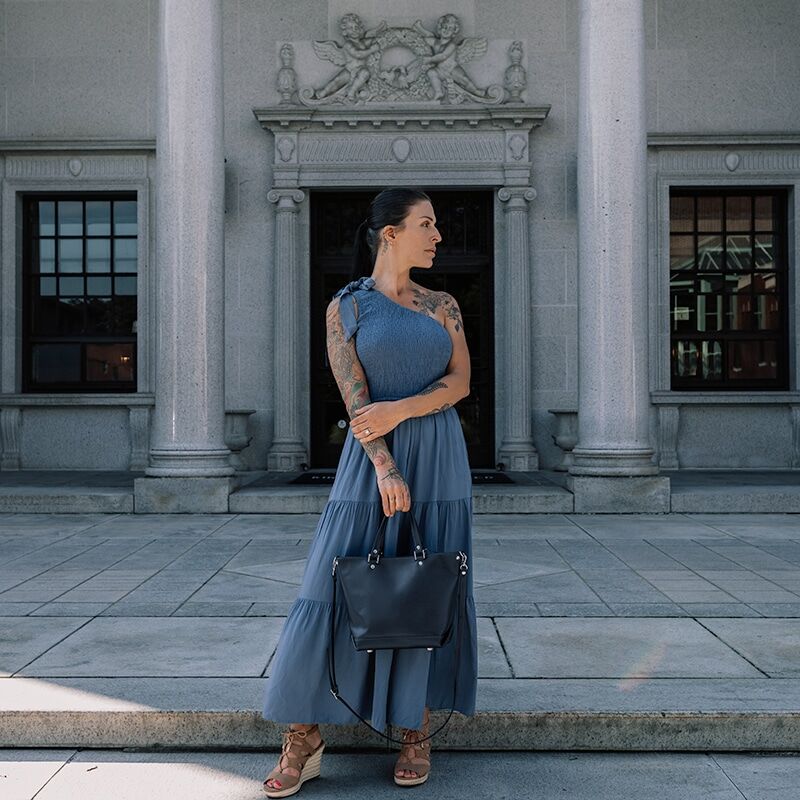 Woman in blue dress carrying a navy blue leather purse in front of a building with columns.