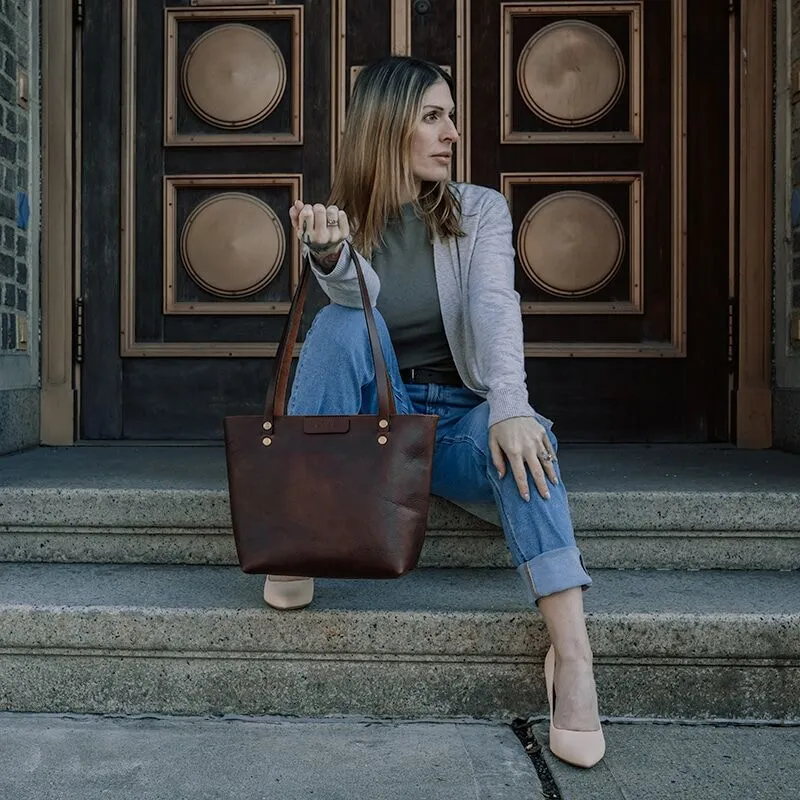 Woman sitting on a set of outside stairs holds a brown leather shoulder bag on her wrist.