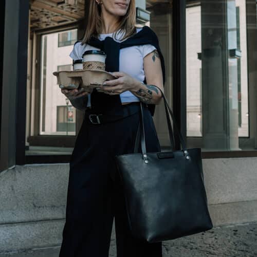 Woman carrying a to-go carrier of coffee with a large black, leather shoulder bag on her arm on her way to work.