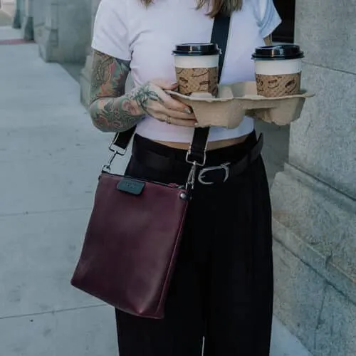 Woman wearing work attire while also wearing a sangria purple leather crossbody bag and carrying two coffees in a carrier.