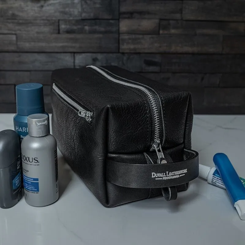 A black leather toiletry bag or Dopp kit sitting on a white marble bathroom counter along with a few toiletries.