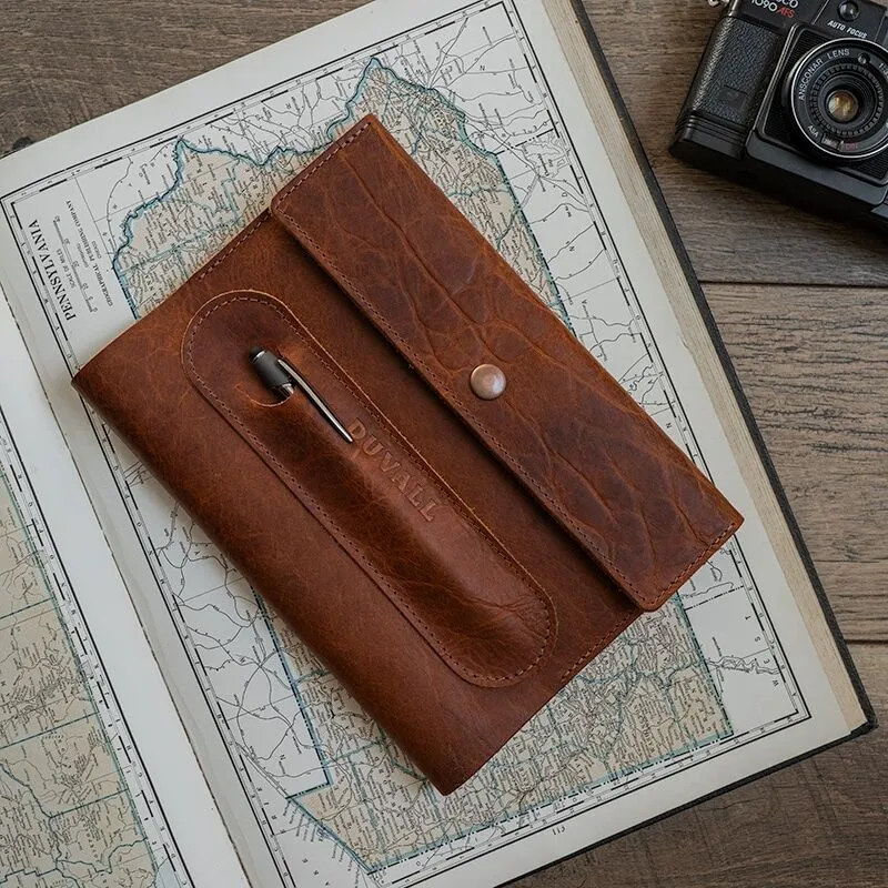 Exterior cover of the brown bison leather Journal Cover from Duvall Leather that features an exterior pen pocket stamped with "Duvall", laying on top of an atlas on a wood surface with a camera next to it.