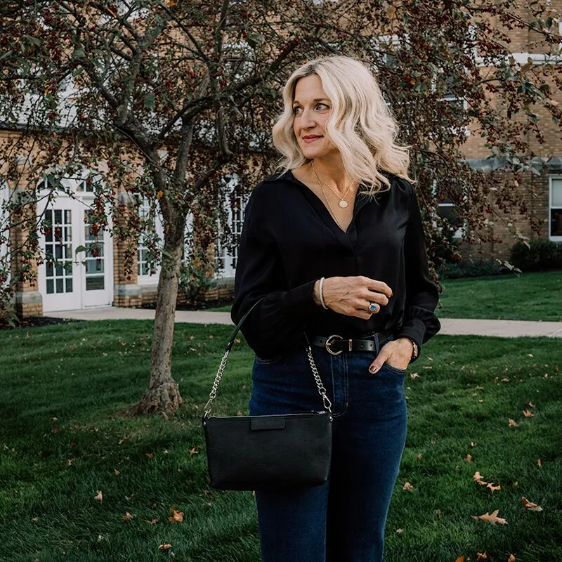A woman wearing a black long sleeved blouse and dark blue jeans carries a small black classy leather handbag that has a part leather/part chain shoulder strap.