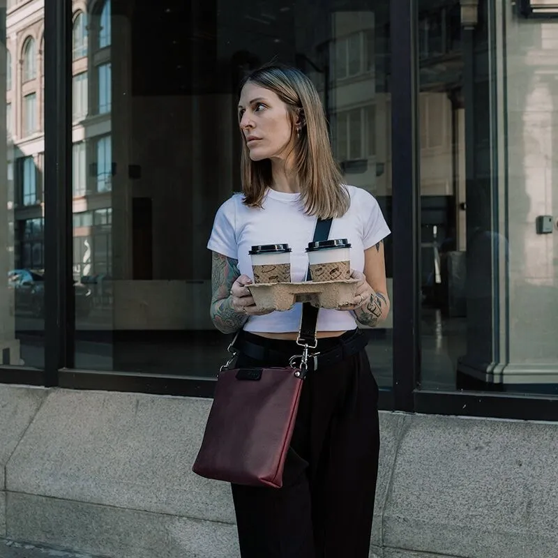 A sangria-purple leather crossbody bag with a black crossbody strap worn by a woman holding a to-go carrier of coffee on her way to work.