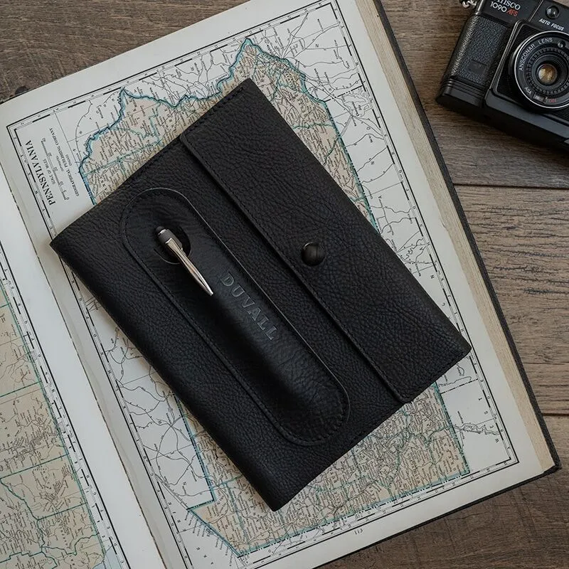 Exterior cover of the black leather Journal Cover from Duvall Leather that features an exterior pen pocket stamped with "Duvall", laying on top of an atlas on a wood surface with a camera next to it.