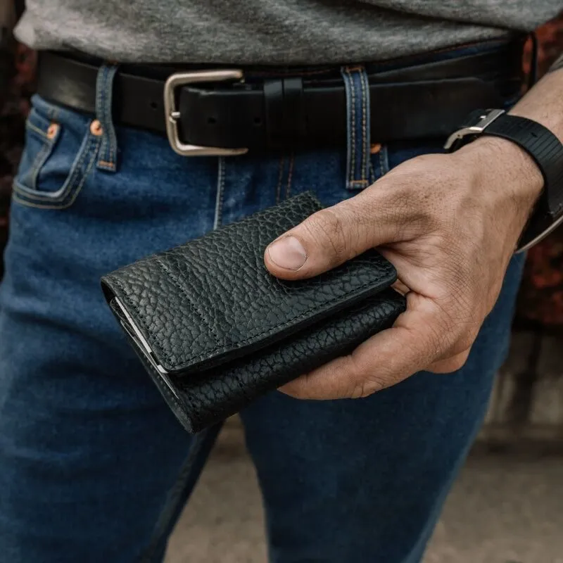 Man holding a closed black bison leather trifold wallet in his hand.