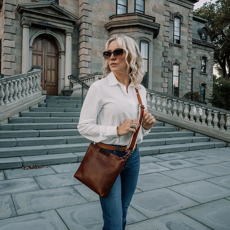Woman with sunglasses wearing a white button down shirt and blue jeans is sporting a red brown leather crossbody bag.