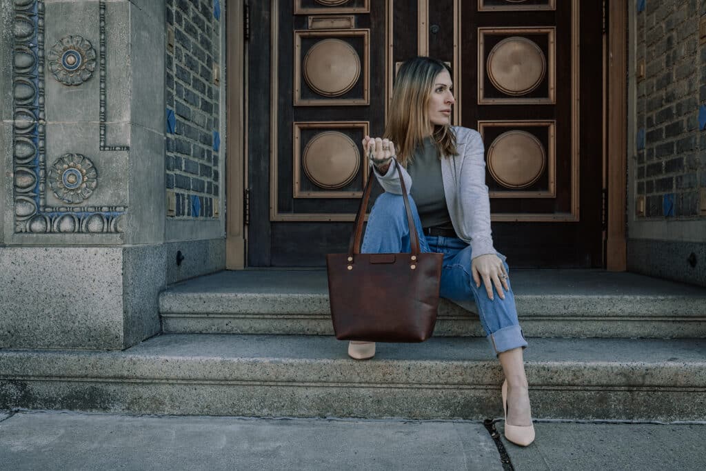 Woman sitting on outdoor set of stairs outside of a brick building with a red brown leather shoulder bag from Duvall Leather.
