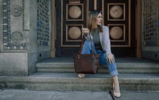 Woman sitting on outdoor set of stairs outside of a brick building with a red brown leather shoulder bag from Duvall Leather.