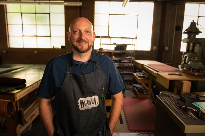 owner of duvall leatherwork, Nick Duvall, standing in his workshop smiling. He is wearing an apron with the Duvall Leatherwork logo