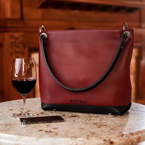 A crimson red and black leather purse is displayed on a marble table. There's a glass of dark red wine nearby.