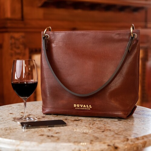 A brown leather purse is displayed on a mable cafe table with a glass of wine and some perosnal accessories
