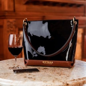A shiny, large black leather bag is displayed on a marble cafe table with a glass of wine
