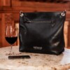 A black leather women's slouch bag displayed to reveal exterior zipper, shown on a marble cafe table with red wine