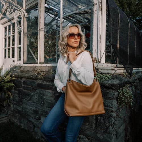 Woman wearing a white button down shirt and blue jeans is leaning up against the wall of a greenhouse while holding a saddle brown leather slouch handbag.