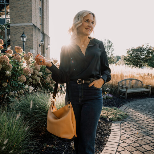 A woman wearing a black blouse and jeans holding a honey and black leather slouch bag during golden hour.
