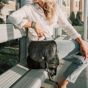 woman sitting on bench wearing a white shirt and blue jeans holding black leather backpack purse