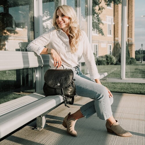 model sitting on a bench holding a black leather backpack purse and wearing a white shirt with blue jeans