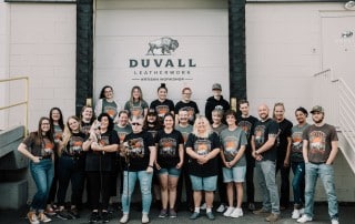 Duvall Leatherwork team photo 2022, the team is standing in front of the warehouse bay door with the duvall leatherwork logo above