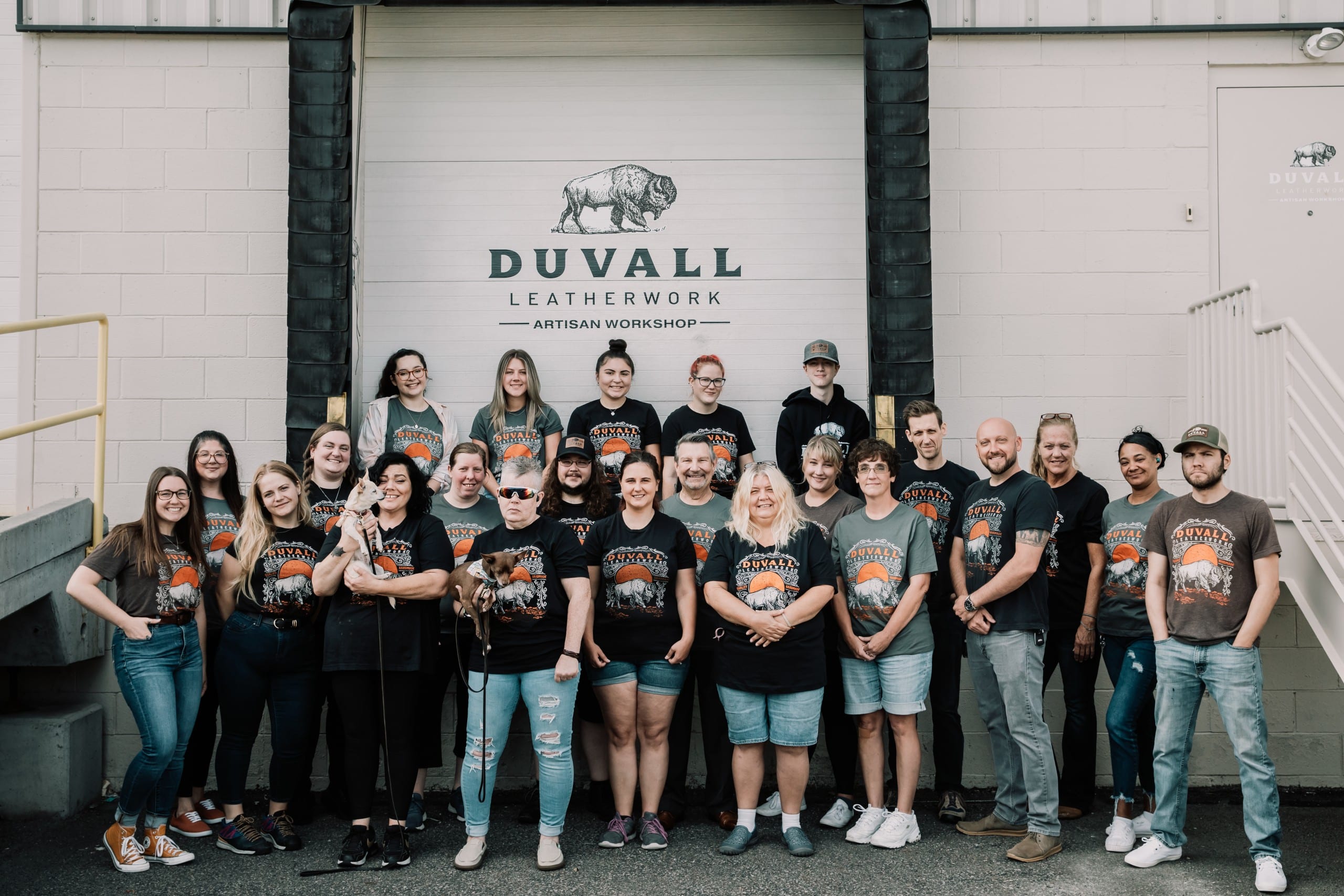 Duvall Leatherwork team photo 2022, the team is standing in front of the warehouse bay door with the duvall leatherwork logo above
