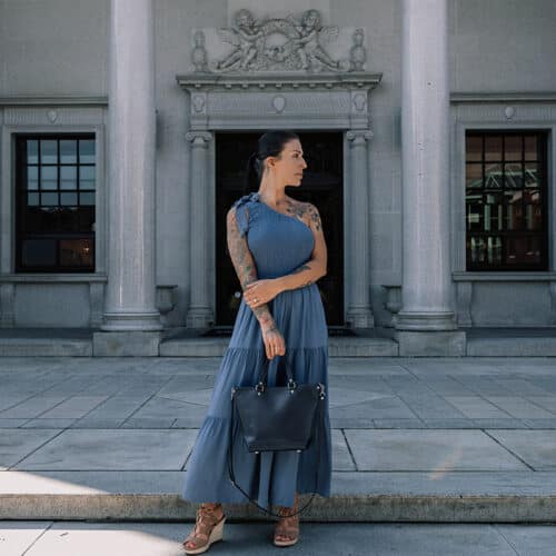 Woman in blue dress carrying a navy blue leather purse in front of a building with columns.