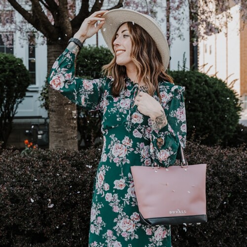 A lovely woman tipping her hat in a green floral dress with a cherry blossom pink purse on her arm with blossom petals falling from the tree.