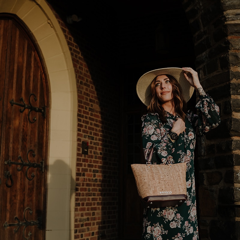 outside shot of model standing in a brick archway, she holds a handbag on one arm and reaches for her hat with the other hand