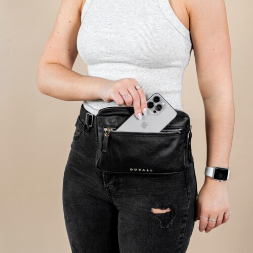 Model in studio wearing black fanny pack pulling out iphone from front zippered pocket.