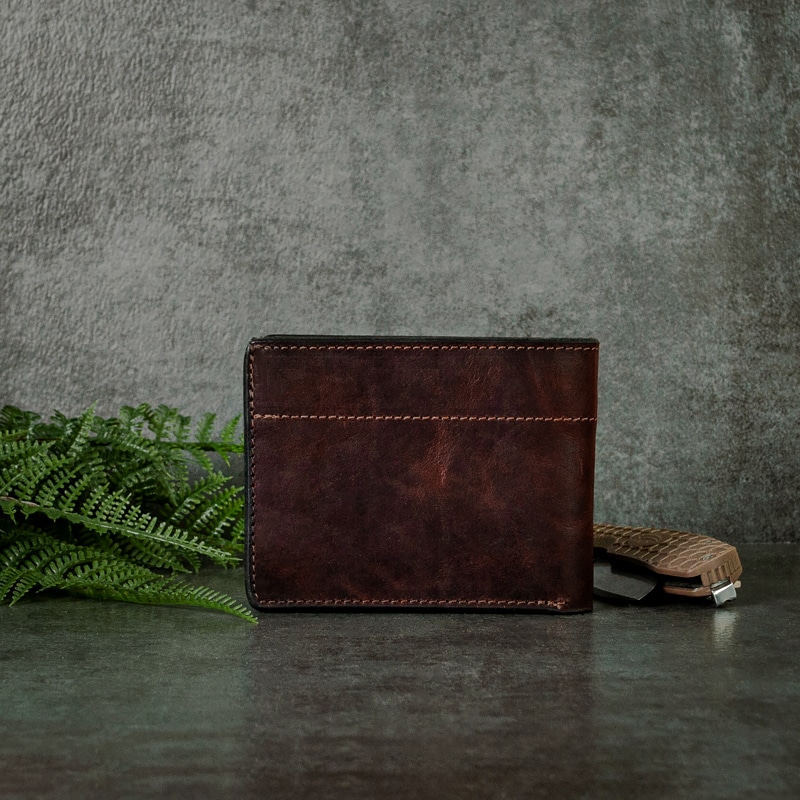 A deep red mahogany bifold wallet that has embroidered stitching around the edges on a slate tabletop background with greenery.