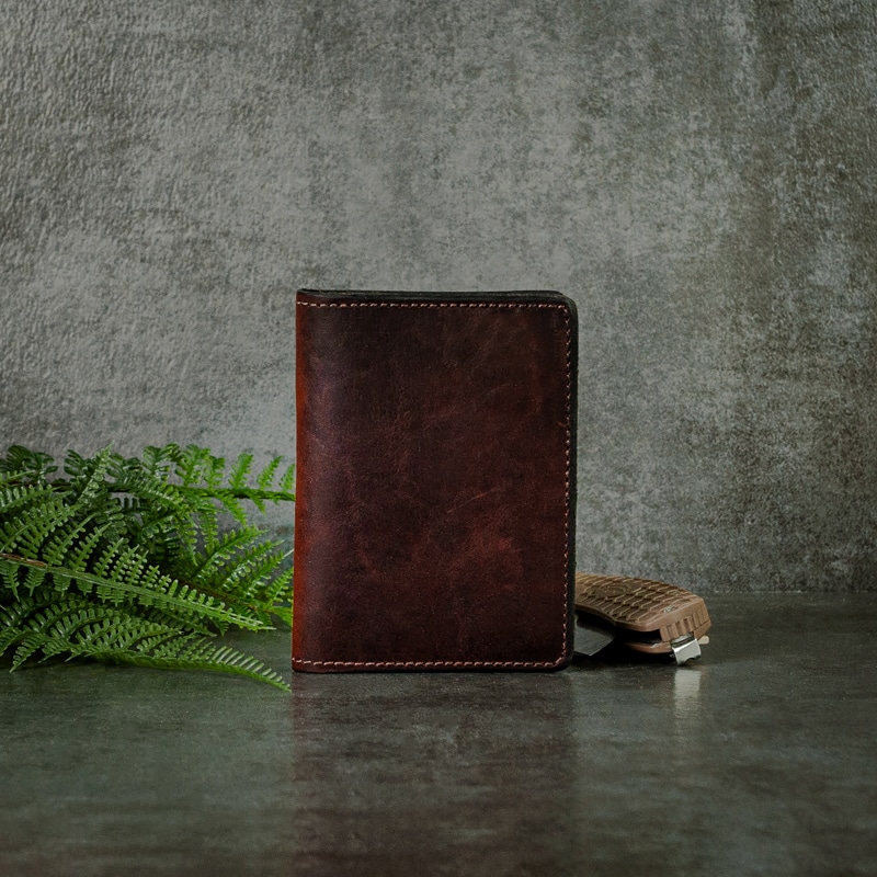 Real dark mahogany leather credit card wallet with fine stitching on the edges photographed with greenery and pocket knife on a slate background.