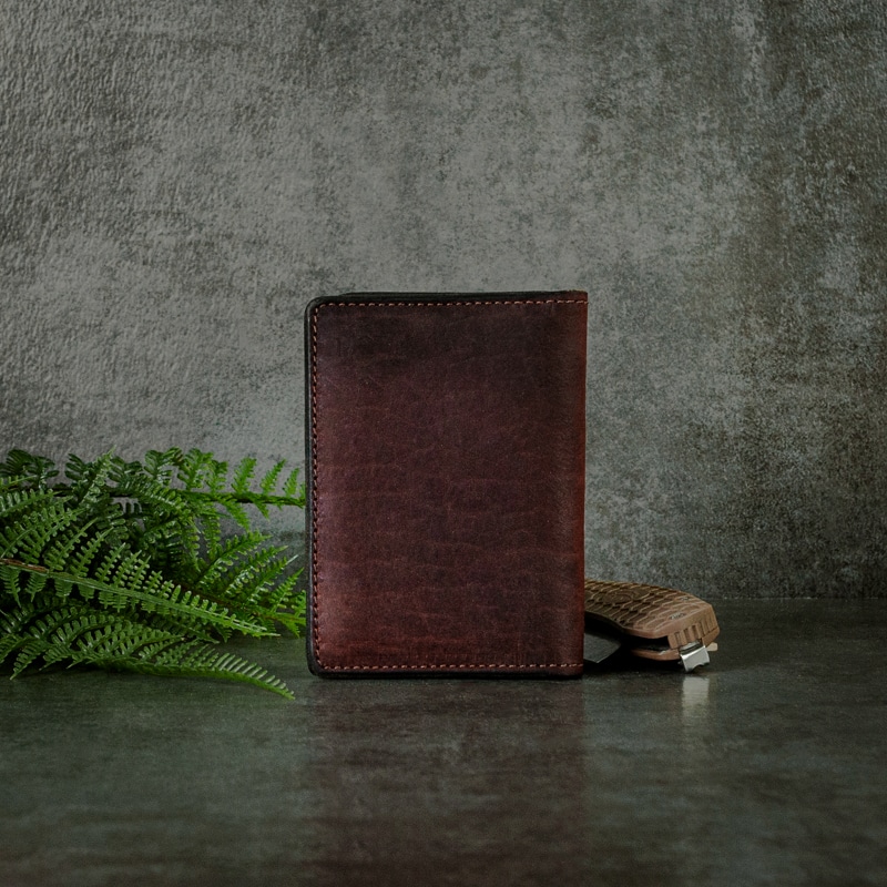 Dark red credit card wallet that has fine stitching on the side that's a great size for his every day usage photographed on slate background with ferns and pocket knife.