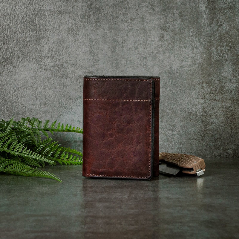Deep mahogany leather trifold wallet that's very masculine shot on a slate background with greenery and a pocket knife.