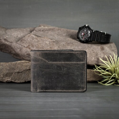Sturdy anthracite goatskin leather bifold wallet is the perfect gift for him for any occasion photographed on grey wood with watch on rocks and greenery.