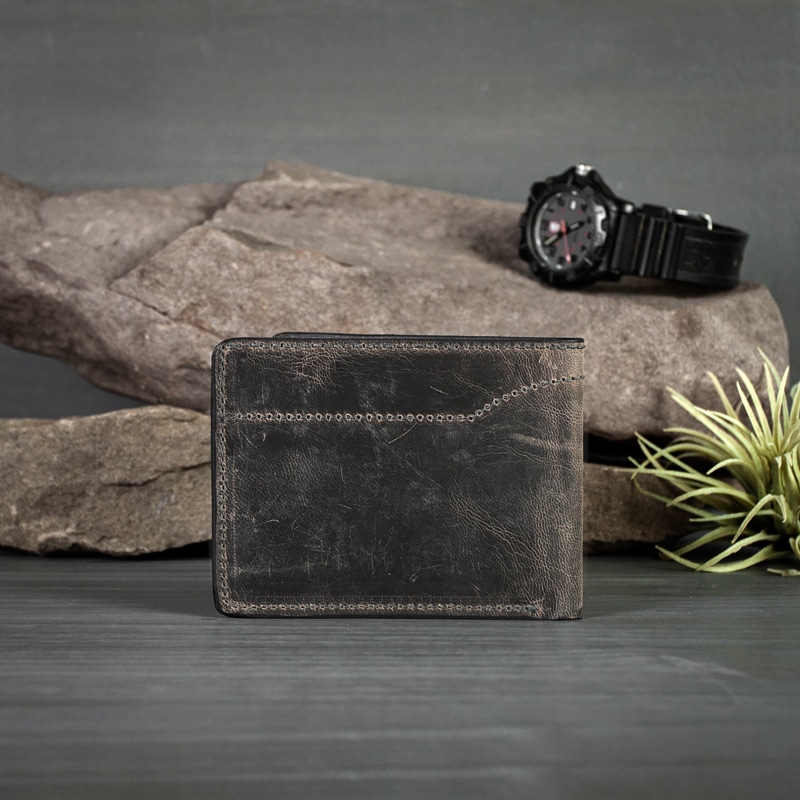 A men's anthracite bifold wallet with fine stitching is hand made in America shot on grey wood with watch on rocks and greenery.
