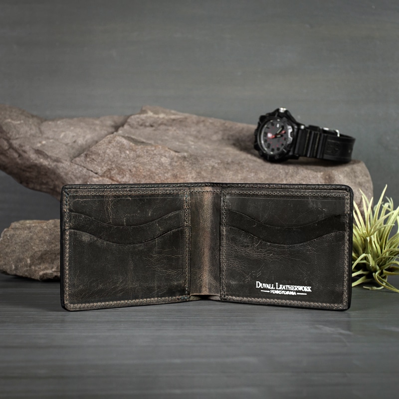 Inside of an anthracite bifold wallet that holds multiple card holders with inner pockets for more storage on grey wood with a watch on rocks and greenery.