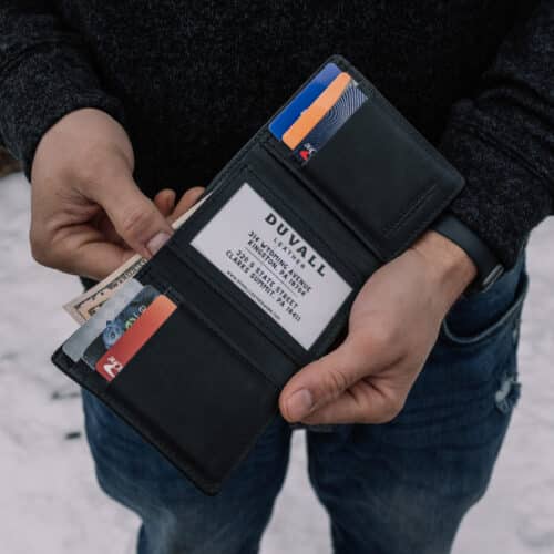 Red Bison Trifold Wallet opened in a man's hands to show the black goatskin leather interior filled with credit cards and cash, also shows the presence of the ID window.