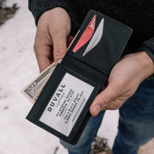 Red Bison Bifold Wallet with ID window opened in a man's hands to show the black goatskin leather interior filled with credit cards and cash, also shows the presence of the ID window.