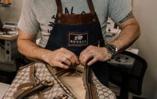 A photograph of the owner of Duvall Leather repairing a designer leather handbag by sewing a new zipper onto the overnight bag.