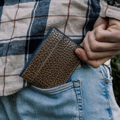 A man taking a men's leather green bison bifold wallet out of the back pocket of his light blue denim jeans.