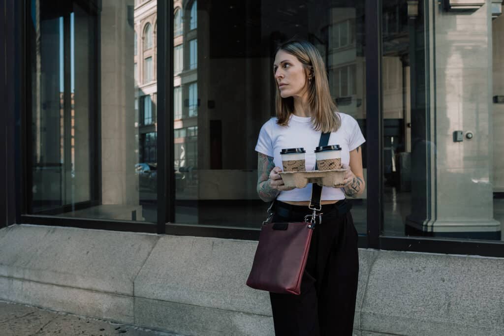 Woman wearing work attire is also wearing a sangria purple leather crossbody bag while also holding a carrier with two coffees.