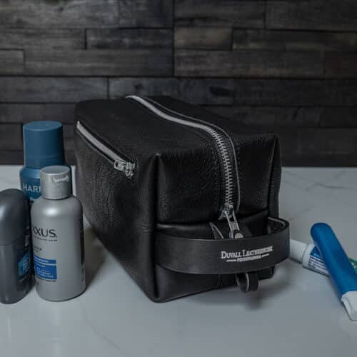 A black leather toiletry bag or Dopp kit sitting on a white marble bathroom counter along with a few toiletries.
