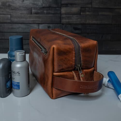 A brown bison leather toiletry bag or Dopp kit sitting on a white marble bathroom counter along with a few toiletries.