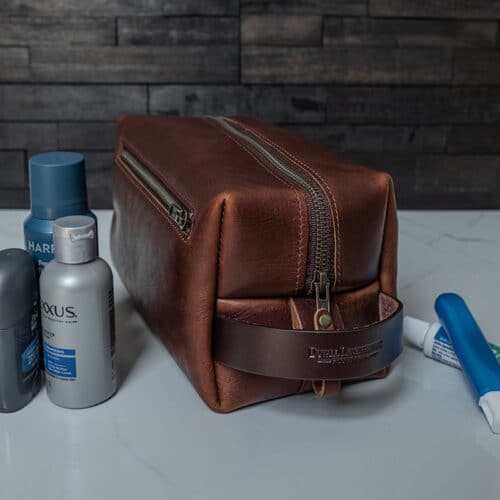 A red brown leather toiletry bag or Dopp kit sitting on a white marble bathroom counter along with a few toiletries.