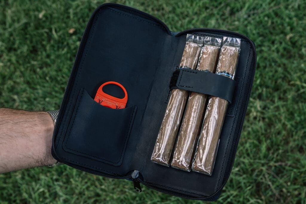 Man wearing a dress watch is holding an open black leather cigar case showing off its ability to hold 3 cigars and a cigar cutter inside.