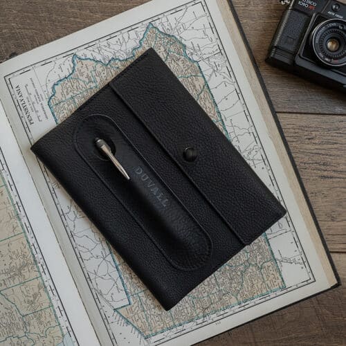 Exterior cover of the black leather Journal Cover from Duvall Leather that features an exterior pen pocket stamped with "Duvall", laying on top of an atlas on a wood surface with a camera next to it.