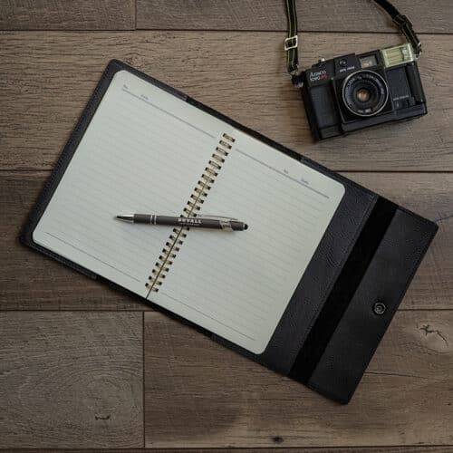 Black Leather Journal Cover with a notebook inside laid out on a wood surface with a gray Duvall Leather pen laying on top of the notebook.