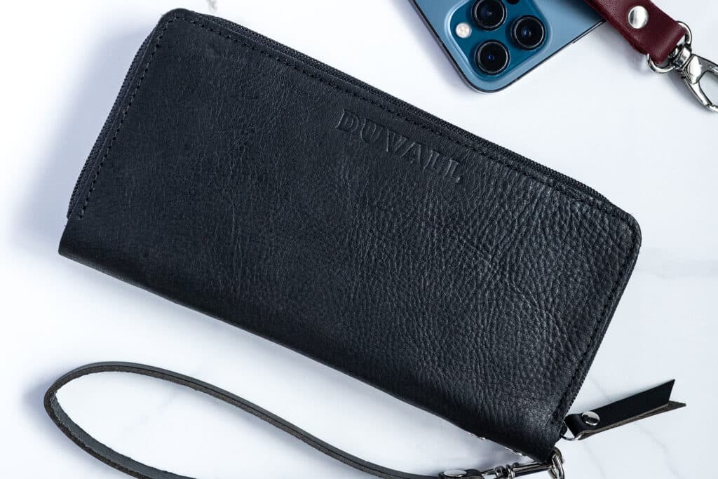 A black Zip-Around wallet laying on a white marble countertop with a blue iPhone and a set of keys