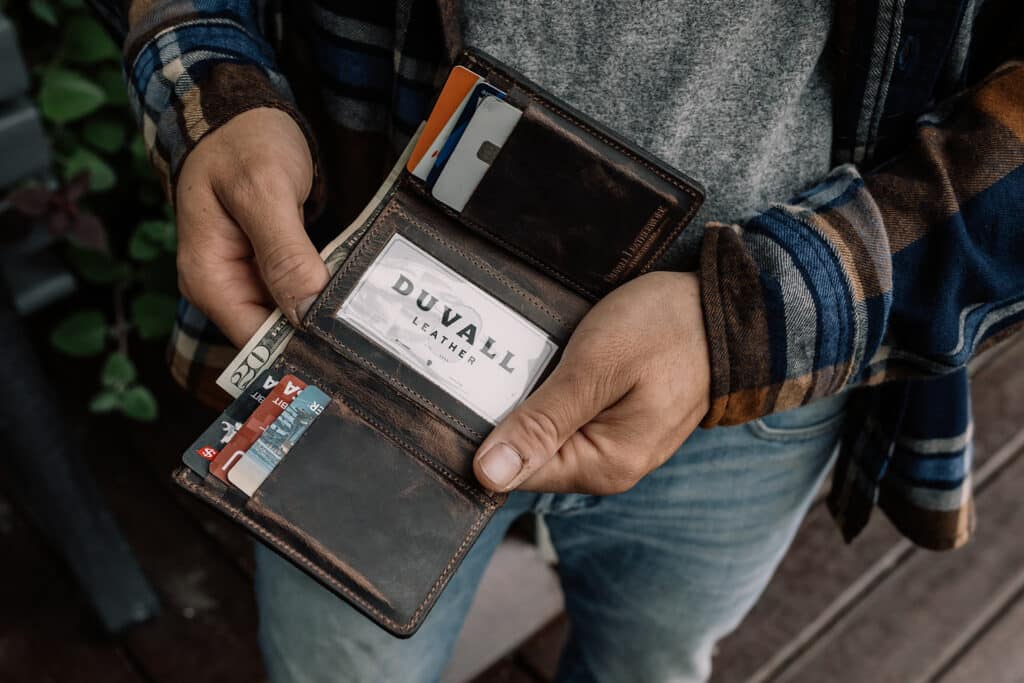 Man holding a chocolate brown leather trifold wallet open to show the card slots and cash inside.