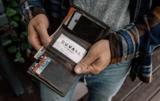 Man holding a chocolate brown leather trifold wallet open to show the card slots and cash inside.