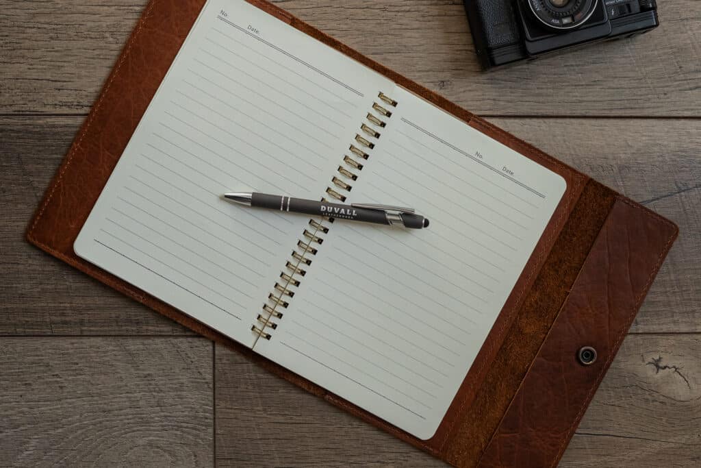 Brown Bison Leather Journal Cover with a notebook inside laid out on a wood surface with a gray Duvall Leather pen laying on top of the notebook.